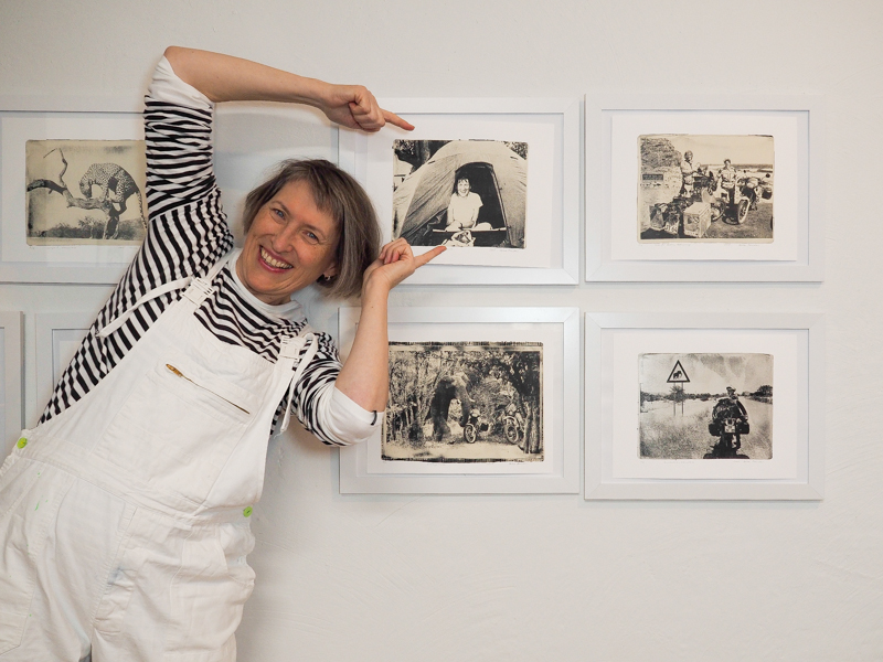 Das Bild zeigt ein Porträt der Künstlerin Heike Pander in ihrem Atelier. Sie steht neben ihren Gel-Drucken an der Wand und zeigt auf die Bilder. Auf ihnen sind Reiseerinnerungen ihrer Motorradreise im südlichen Afrika zu sehen. Alle Bilder sind gedruckte Fotounikate von der Gel-Platte.