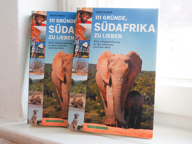 "111 Gründe, Südafrika zu lieben", Autorin Heike Pander Das Bild zeigt ein Foto des Buchs "111 Gründe, Südafrika zu lieben" der Autorin Heike Pander. Zwei Bücher sind zu sehen, die gegen ein Fenster gelehnt sind. Das Cover zeigt die Überschrift, sowie eine große Aufnahme einer Elefantenkuh mit Kalb und an einer Seitenleiste links von oben nach unten das Foto eines lachenden Jungen mit Hut, eine Küstenimpression, eine Hausansicht, ein Foto vom Sagole Big Tree Baobab, den Blick in das Fenster Gottes, zwei Löwen dösen auf der Straße, ein Erdmännchen.